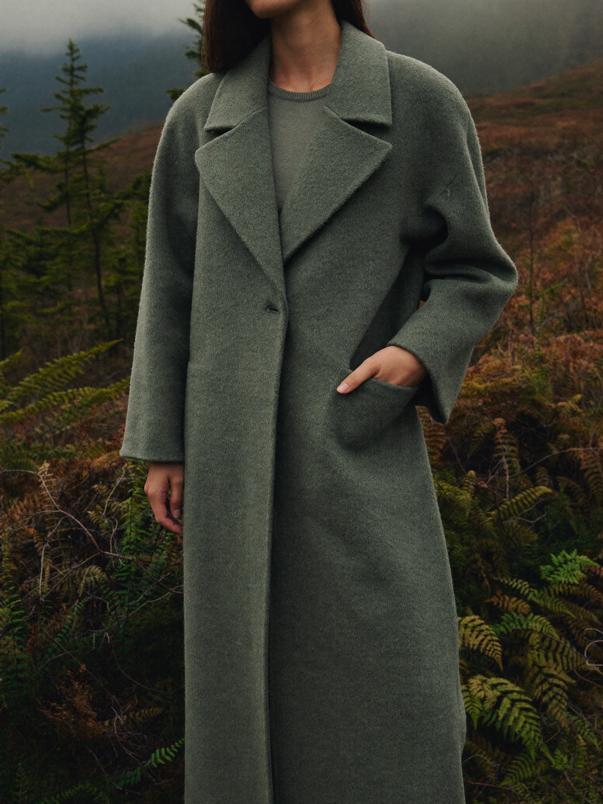 Woman wearing a muted moss green long wool blend overcoat among sword ferns