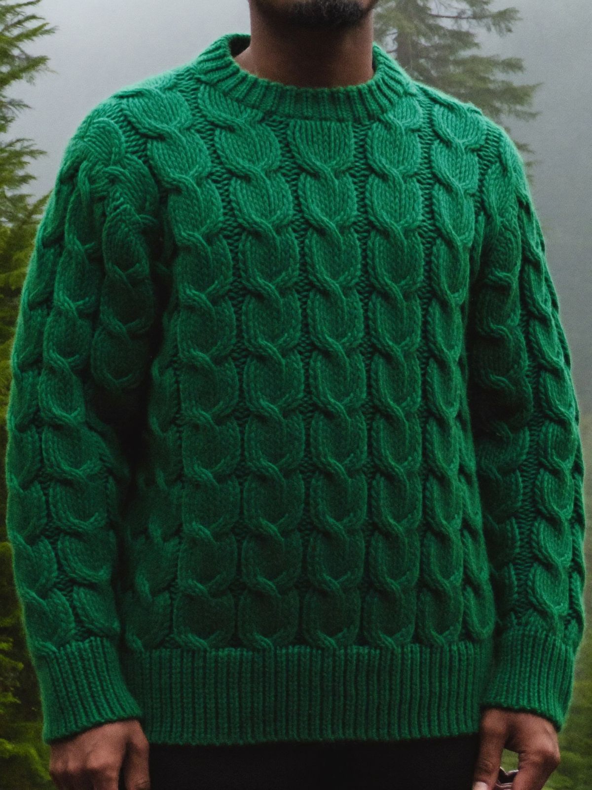Man wearing a deep forest green chunky cable knit wool sweater in a misty forest