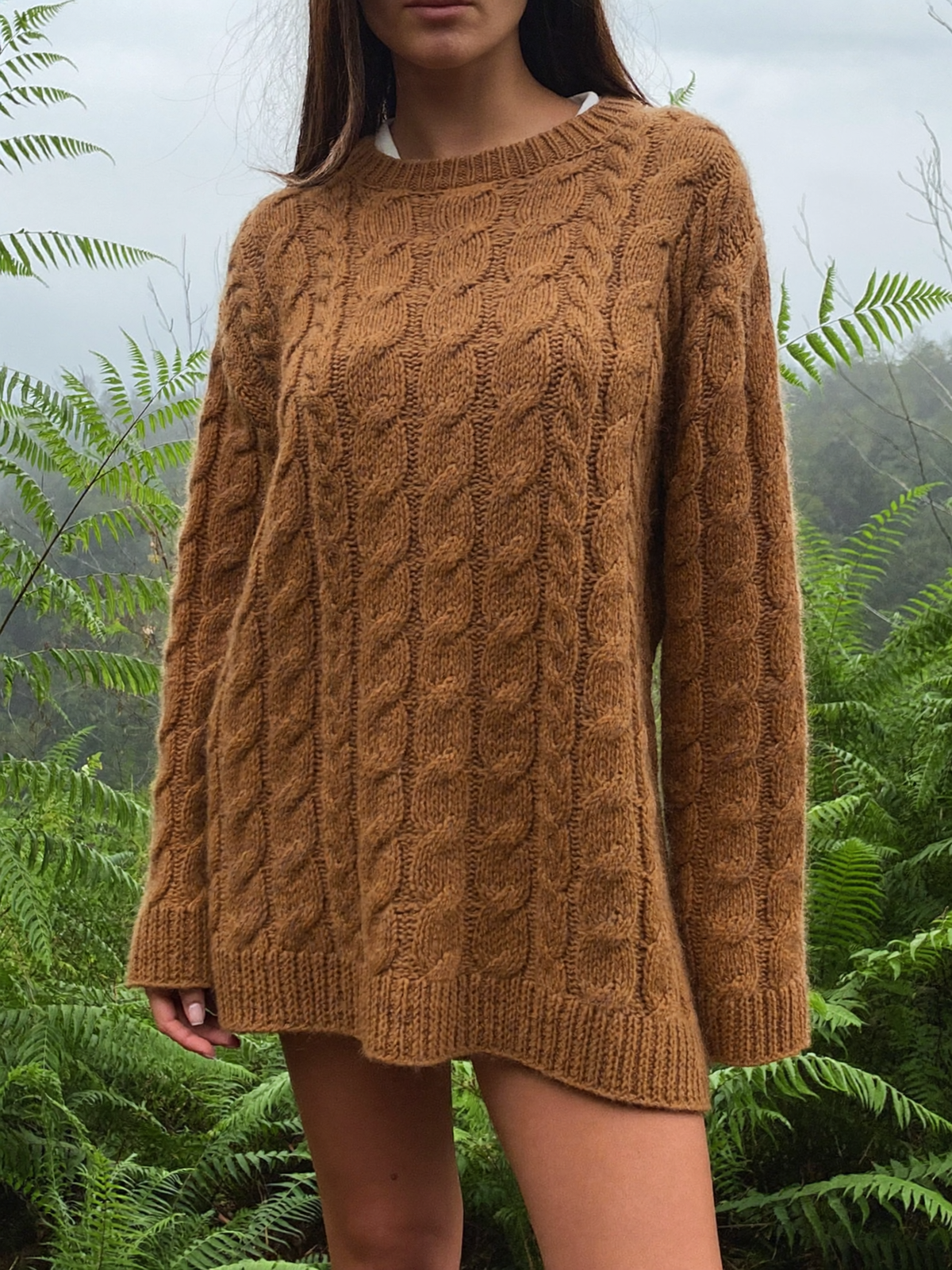 Woman wearing a bark brown merino wool blend cable knit sweater among sword ferns