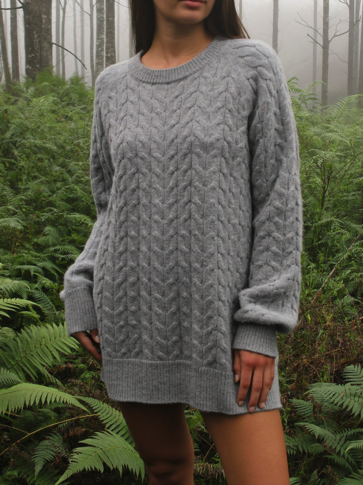 Woman wearing a fog grey merino wool blend cable knit sweater among sword ferns