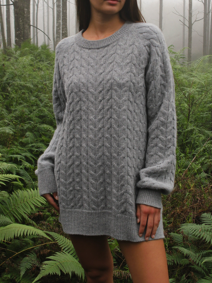 Woman wearing a fog grey merino wool blend cable knit sweater among sword ferns