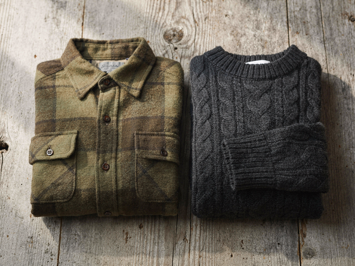 A folded olive and brown brushed flannel shirt beside a charcoal cable-knit wool sweater